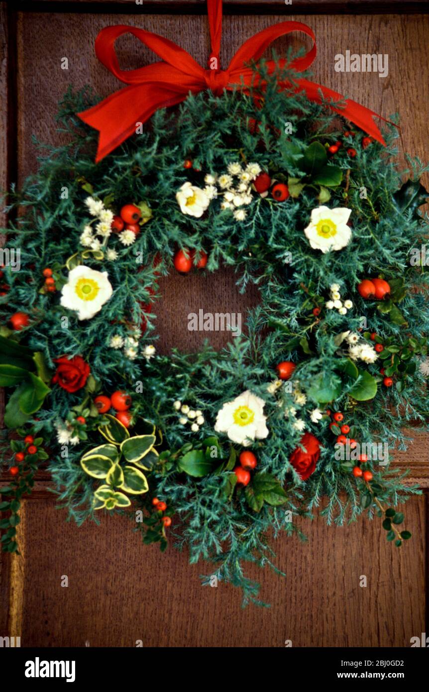 Evergreen christmas wreath with white christmas roses, rosehips, everlasting flowers and dried rosebuds, hanging on oak panelled wall - Stock Photo