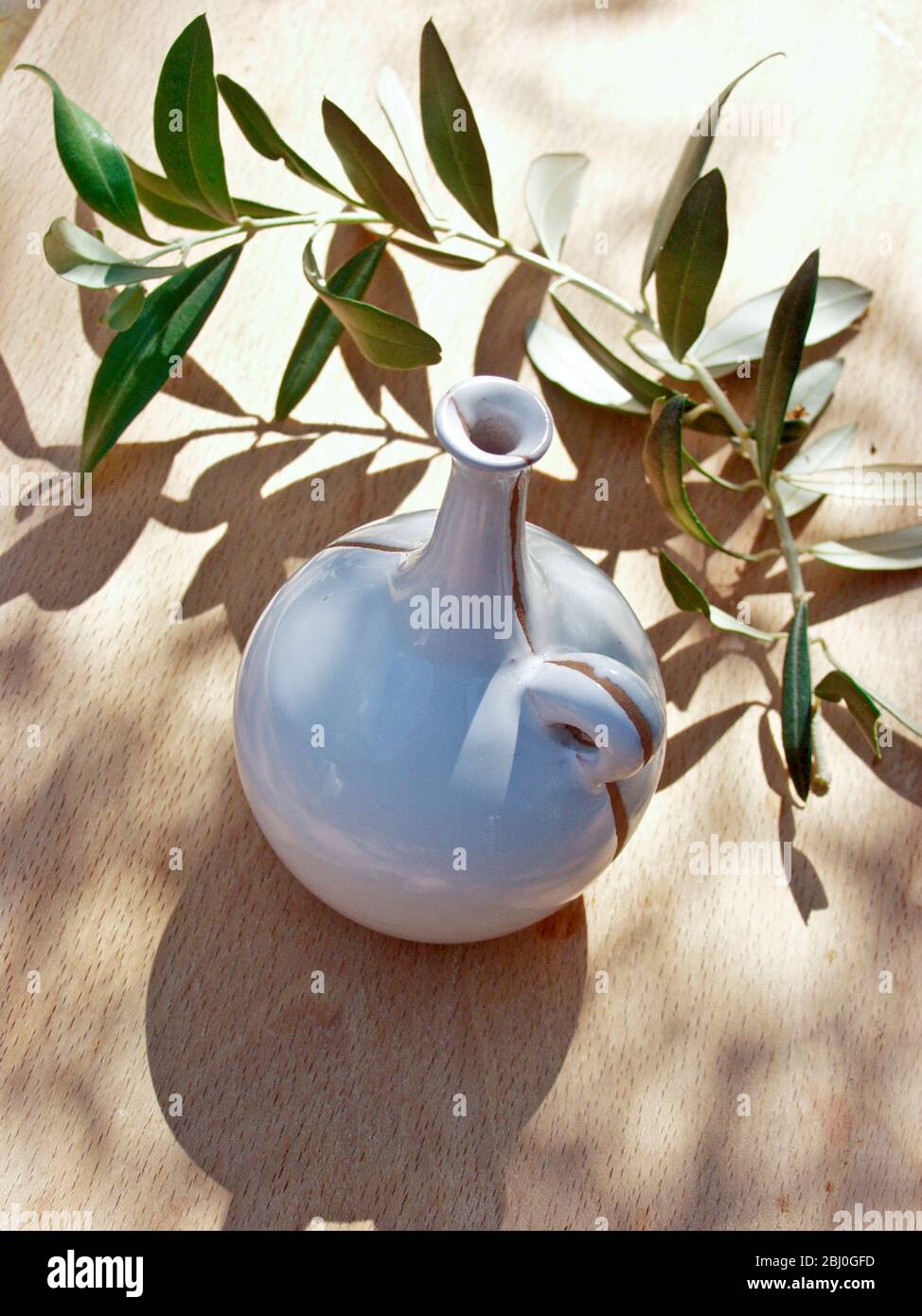 Ceramic oil bottle on tabled dappled in Italian sunshine with branch from olive tree - Stock Photo