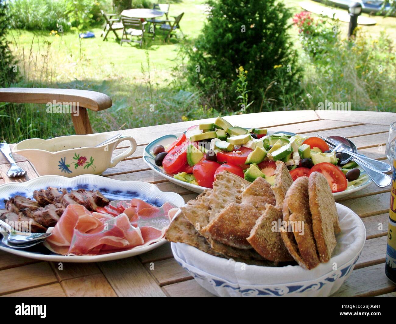 Swedish summer lunch of ham, sausage and cold meat and salad with flat rye bread, served on wooden table in the shade of trees - Stock Photo