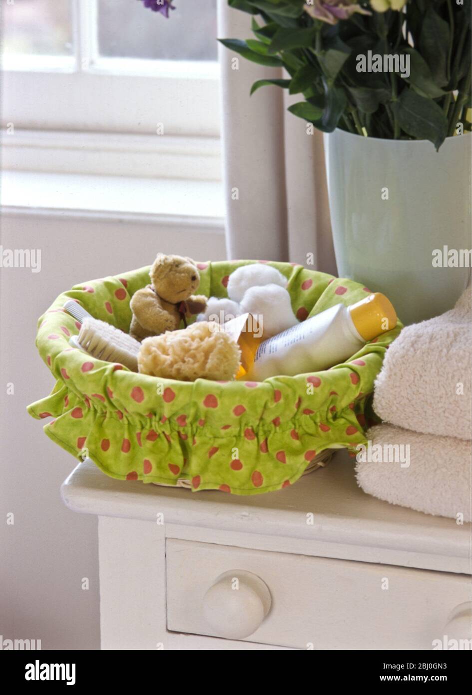 Basket of baby toiletries and little toys, lined with green fabric, in bedroom setting. - Stock Photo