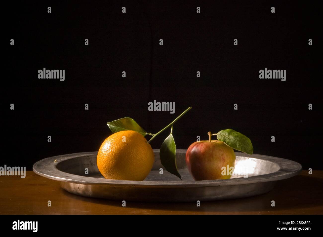 Apple and orange with leaves on pewter charger with black background - Stock Photo