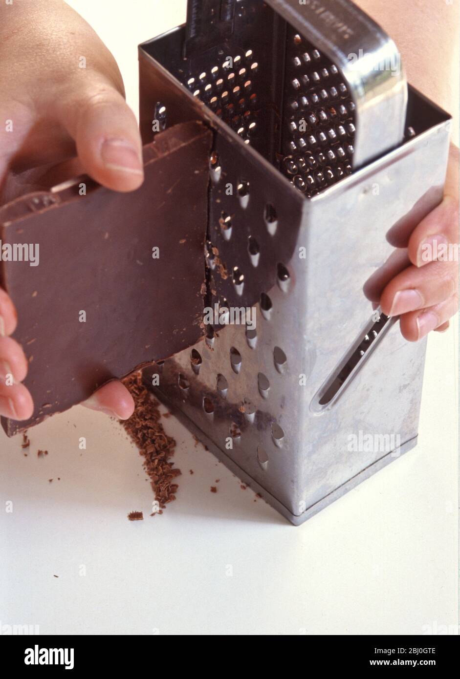 Grating block of dark chocolate on chrome grater. - Stock Photo