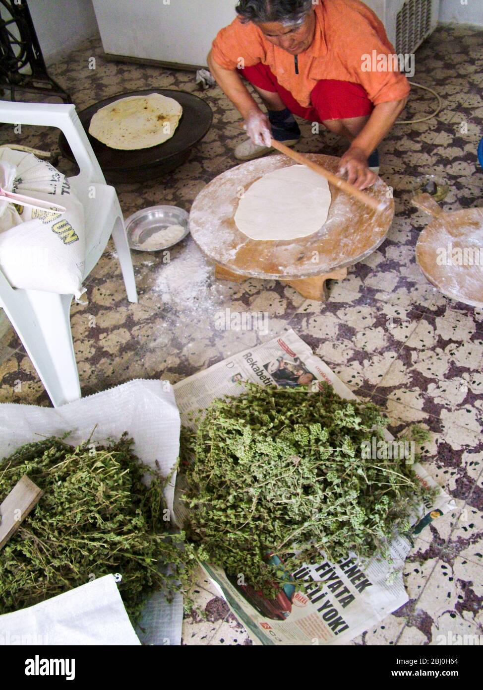 Turkish woman baker rolling out large thin circles of bread on a round wooden board on floor of quayside bakery. Behind is the gas fired hotplate she Stock Photo