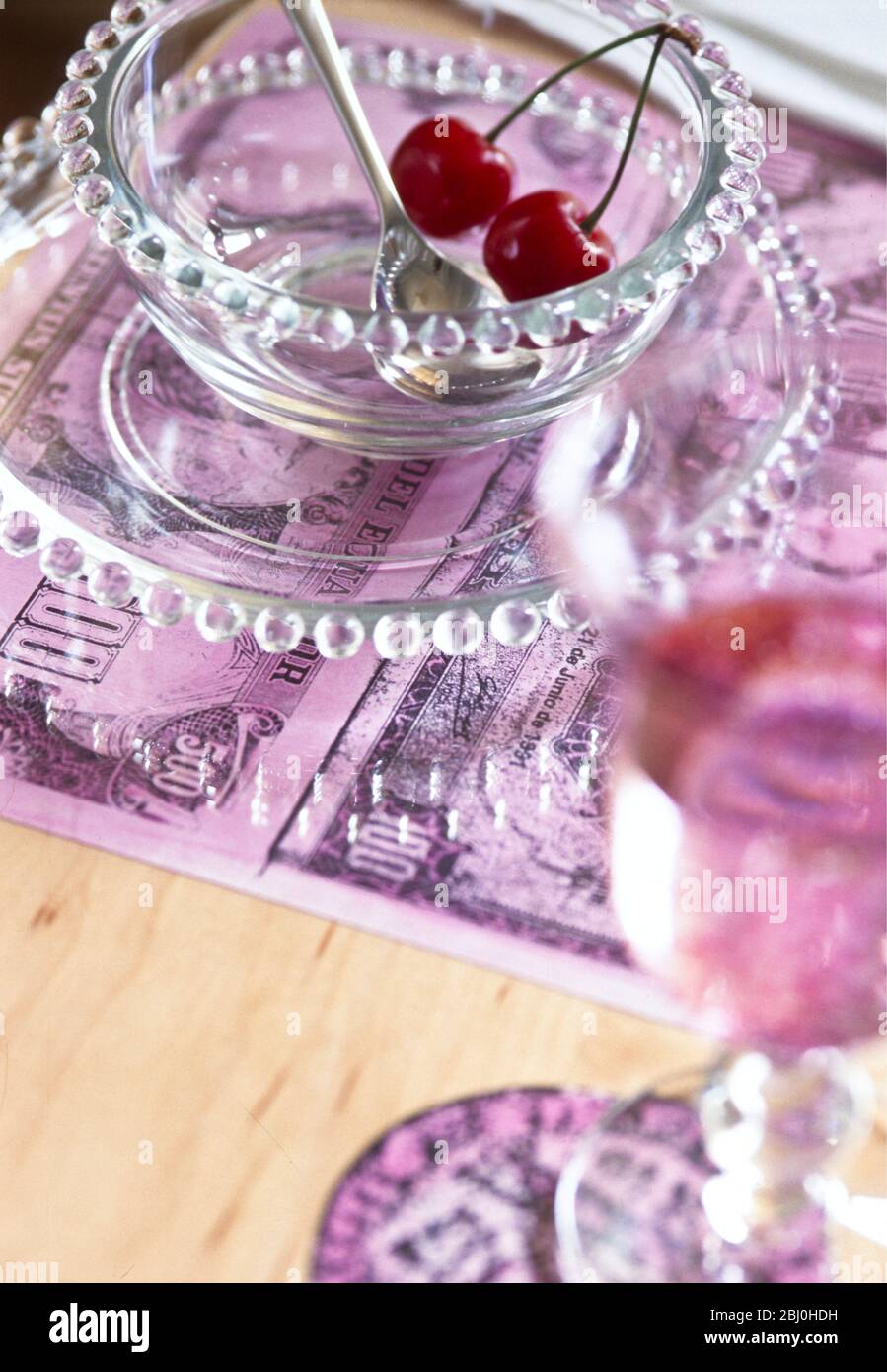 A pair of cherries in a glass bowl with place mat made of enlarged photo copies of old bank notes - Stock Photo
