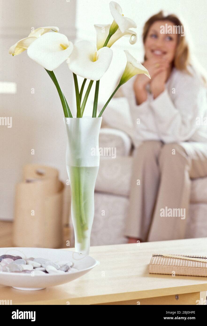 White lilies in tall vase in pale interior with blurred girl on sofa - Stock Photo