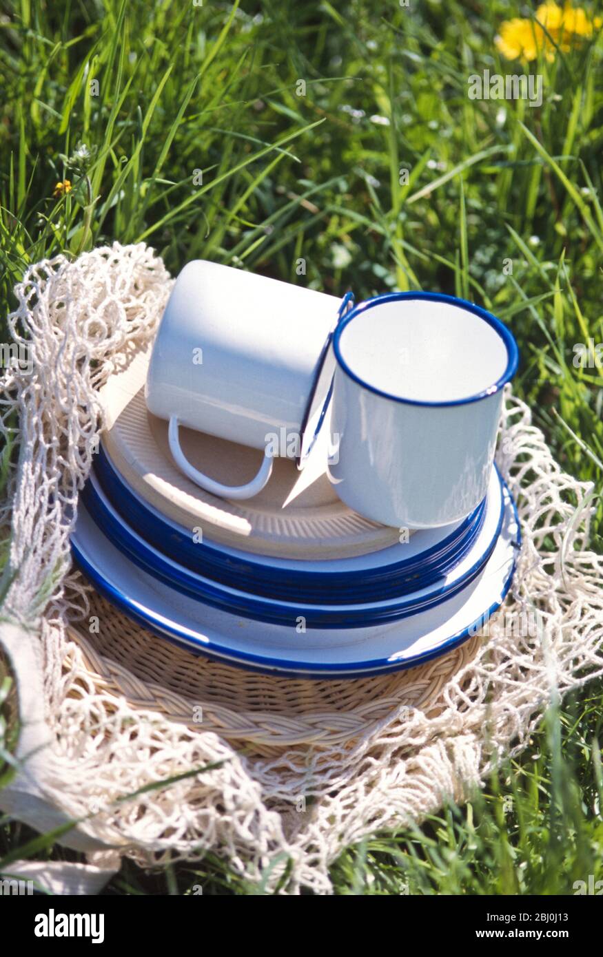 Enamel and paper cups and saucers piled up on a string bag on the grass - part of a spring time picnic - Stock Photo