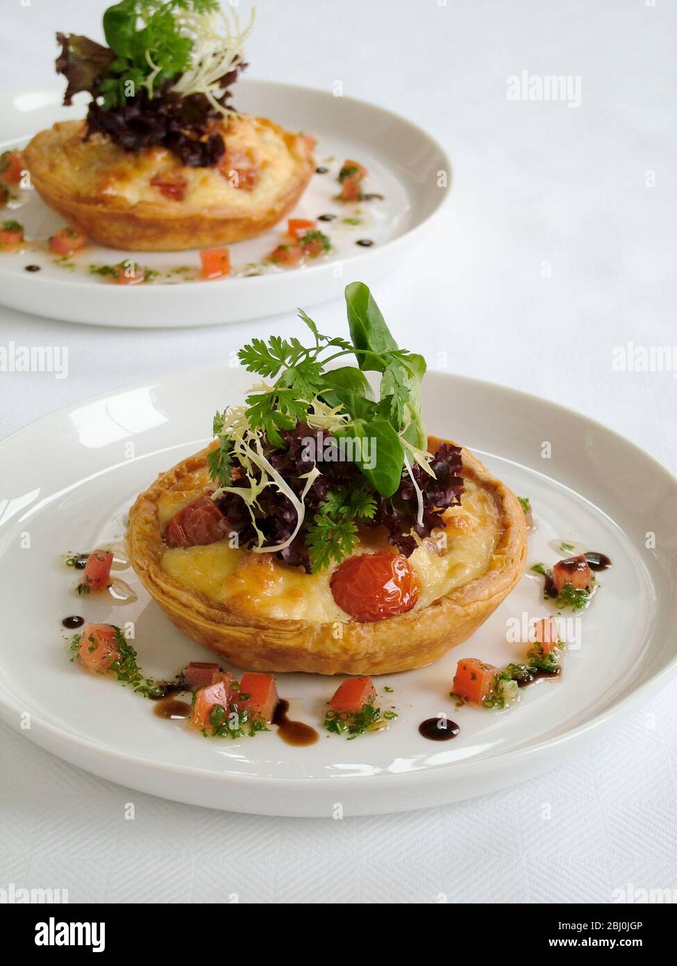 Little savoury party quiches with decorative garnish of baby leaf salad and surrounded by finely chopped tomatoes and herbs with oil and balsamic vine Stock Photo