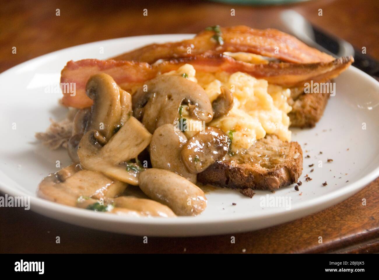 Breakfast plate of mushrooms and scrambled eggs on wholemeal seeded bread with streaky bacon rashers and freshly ground black pepper - Stock Photo