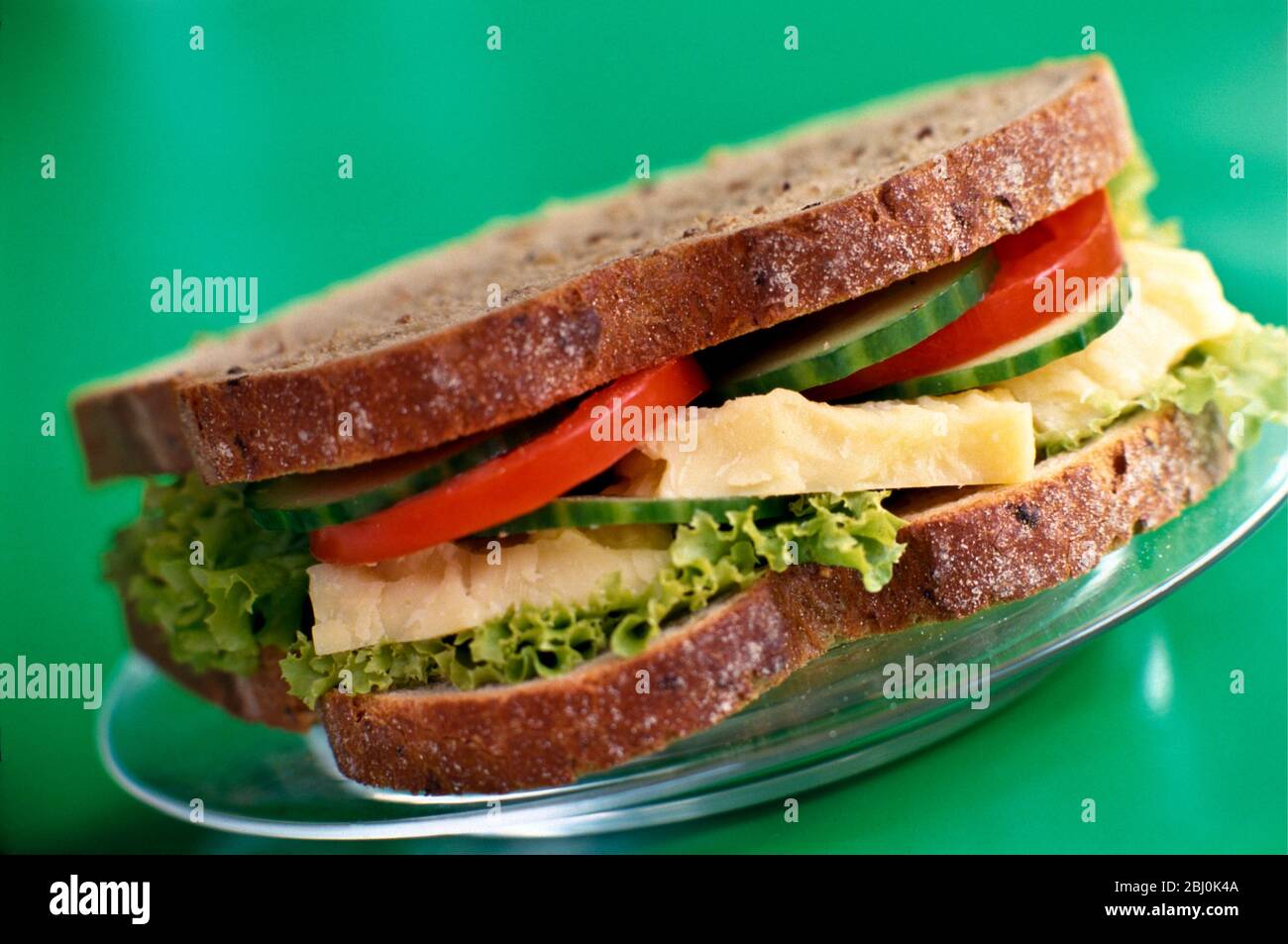Cheese tomato, cucumber and lettuce sandwich on brown wholemeal bread, on glass plate on bright green background - Stock Photo