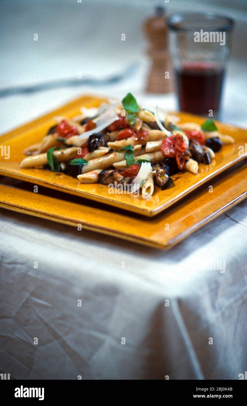 Penne pasta shapes with aubergine, parmesan, basil, tomatoes and olive oil with tumbler of red wine - Stock Photo