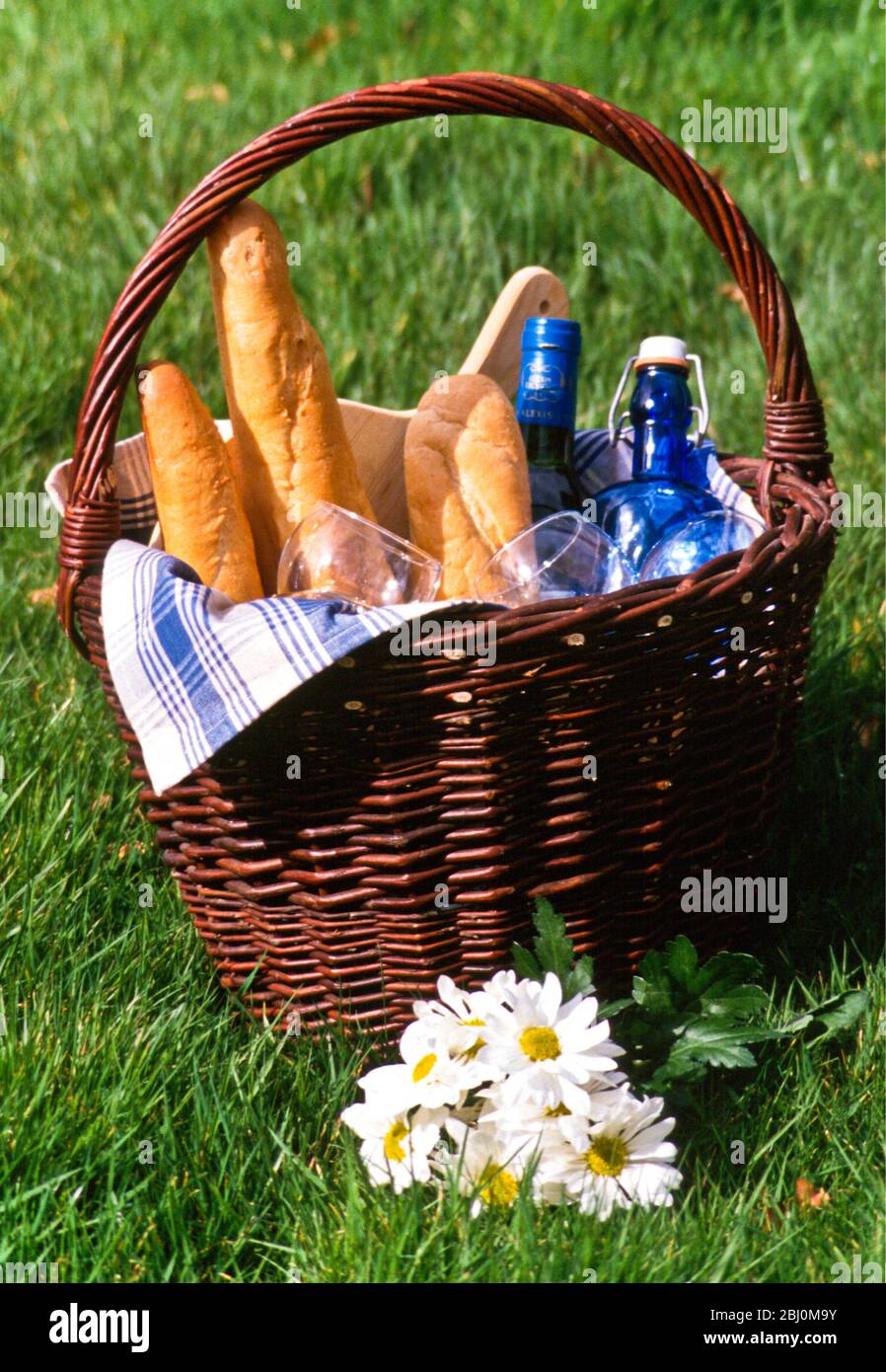 Picnic basket with french bread, glasses and bottles sitting on grass with bunch of daisies - Stock Photo