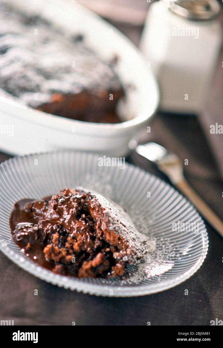 Portion of hot chocolate pudding on glass plate with whole pudding in the background - Stock Photo