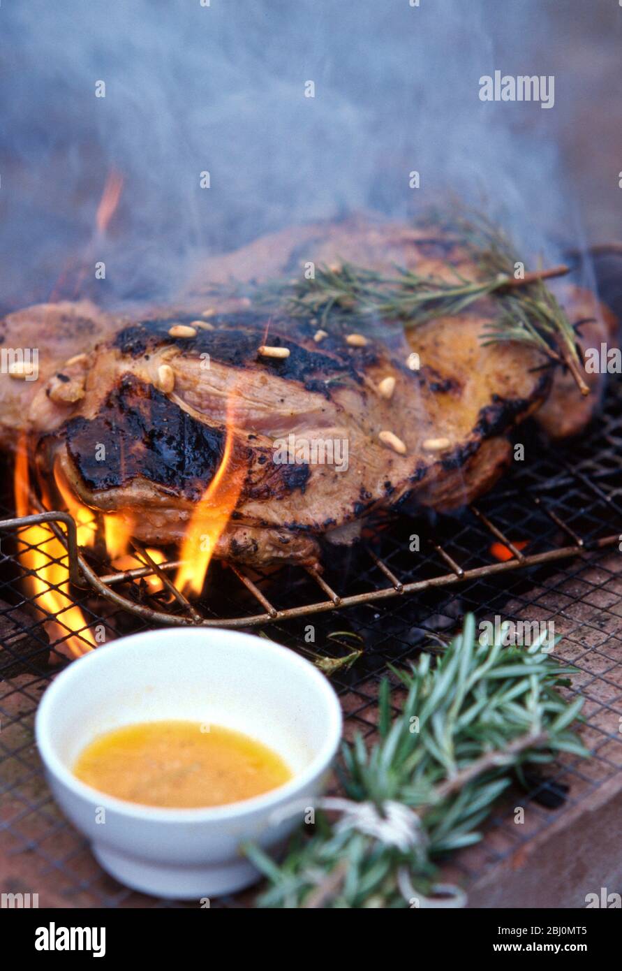 Leg of lamb, butterflied to flatten being barbecued on charcoal grill with rosemary and pine nuts, and 'brush' made of rosemary sprigs used to baste t Stock Photo