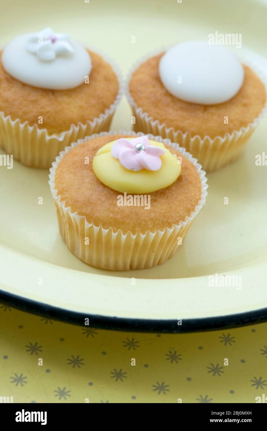 Shop bought fairy cakes on yellow enamel plate - Stock Photo