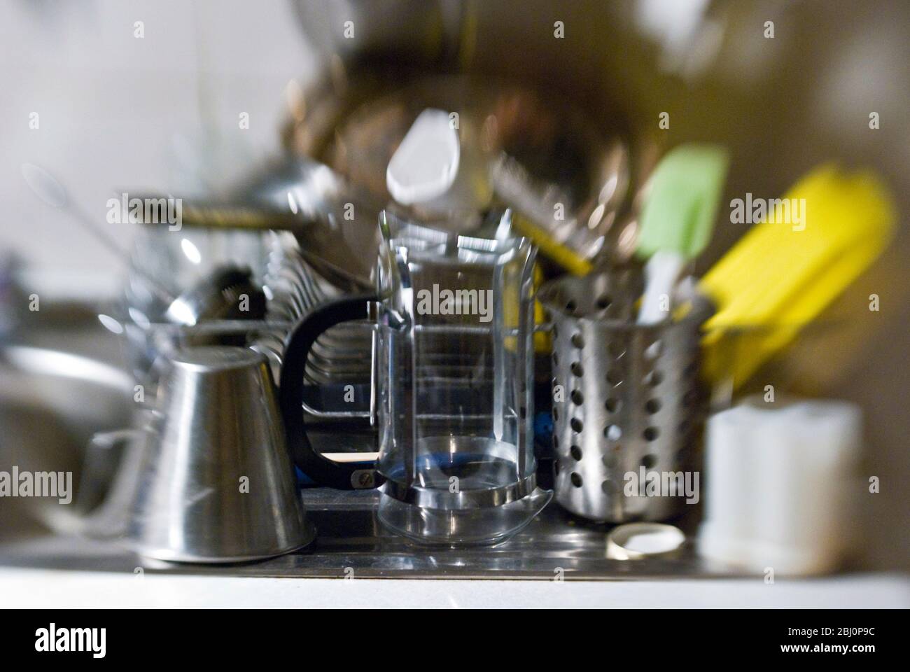 Washing up stacked up in dish drainer on draining board of kitchen sink. Shot with lensbaby lens for blurred effect - Stock Photo