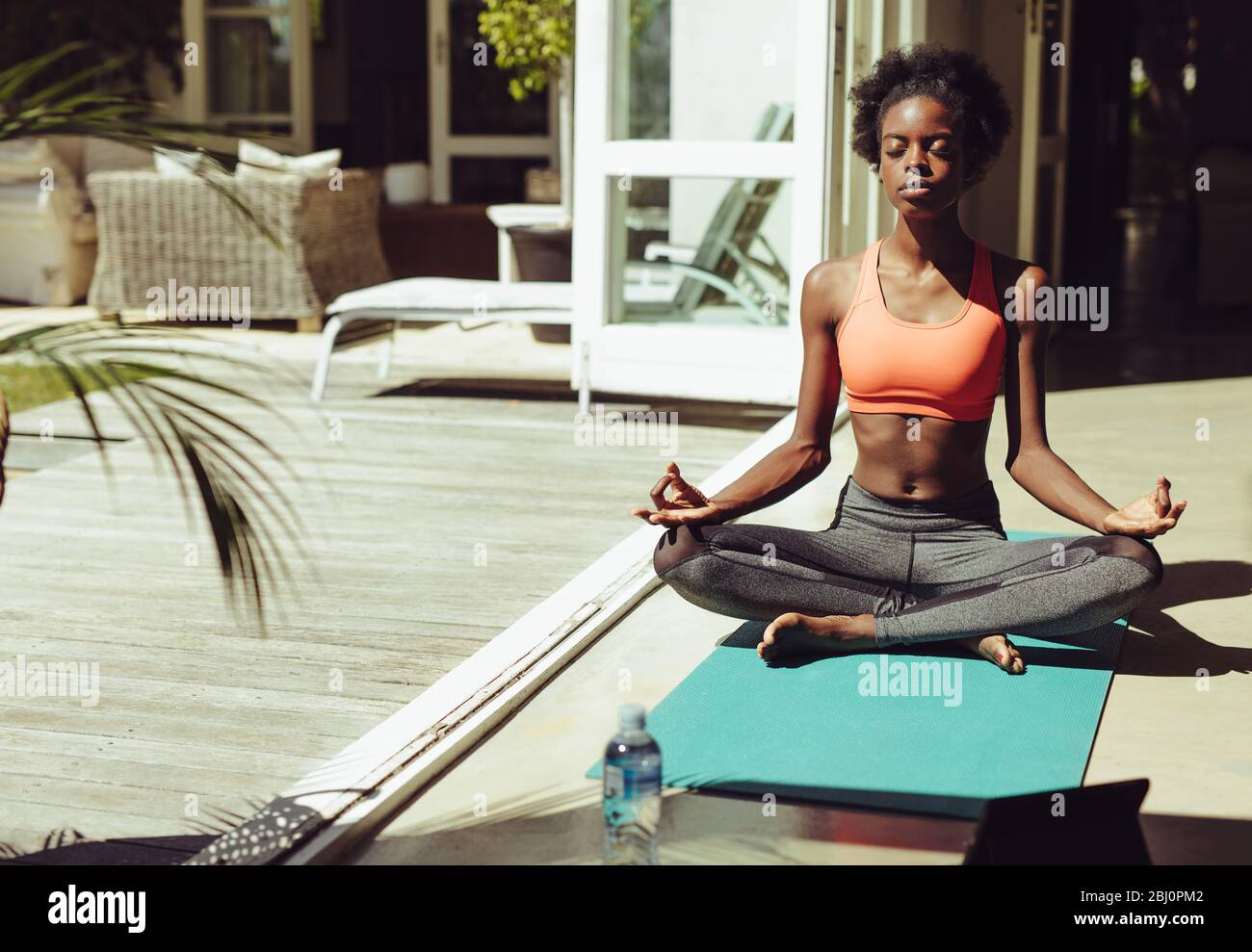 Healthy woman sitting on yoga meditation at home on yoga mat. Fitness woman in sportswear meditating in lotus yoga pose by the poolside. Stock Photo