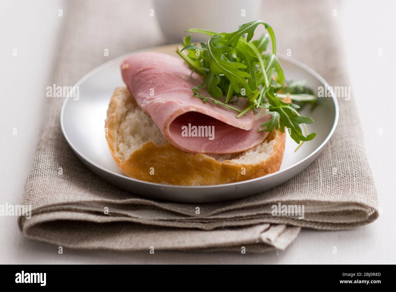 Open sandwich of crusty pain rustique roll, with thick slice of quality ham and rocket salad leaf garnish, with small pot of Dijon mustard - Stock Photo