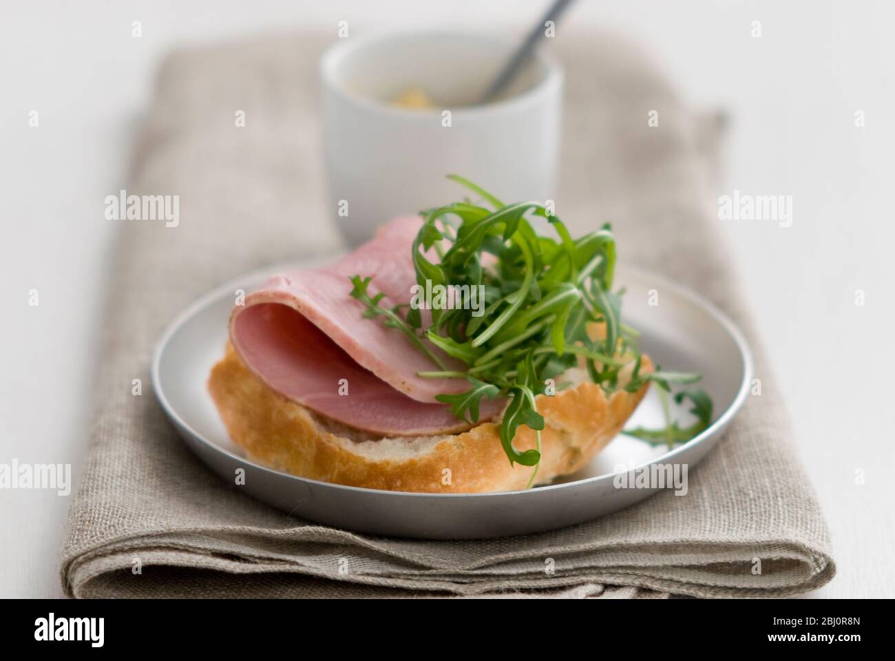 Open sandwich of crusty pain rustique roll, with thick slice of quality ham and rocket salad leaf garnish, with small pot of Dijon mustard - Stock Photo
