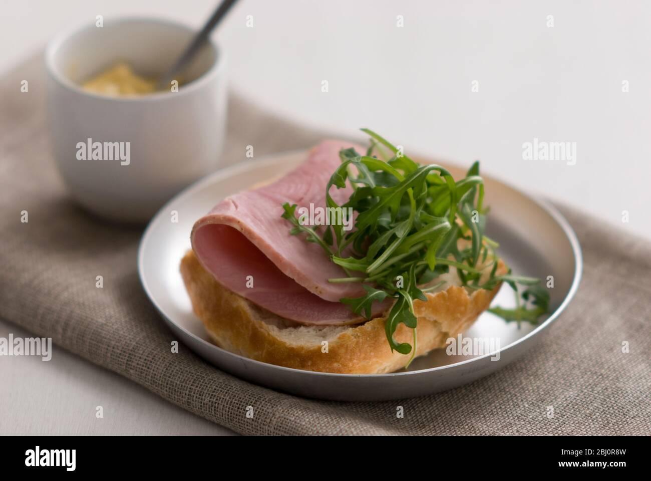 Open sandwich of crusty pain rustique roll, with thick slice of quality ham and rocket salad leaf garnish, with small pot of Dijon mustard - Stock Photo