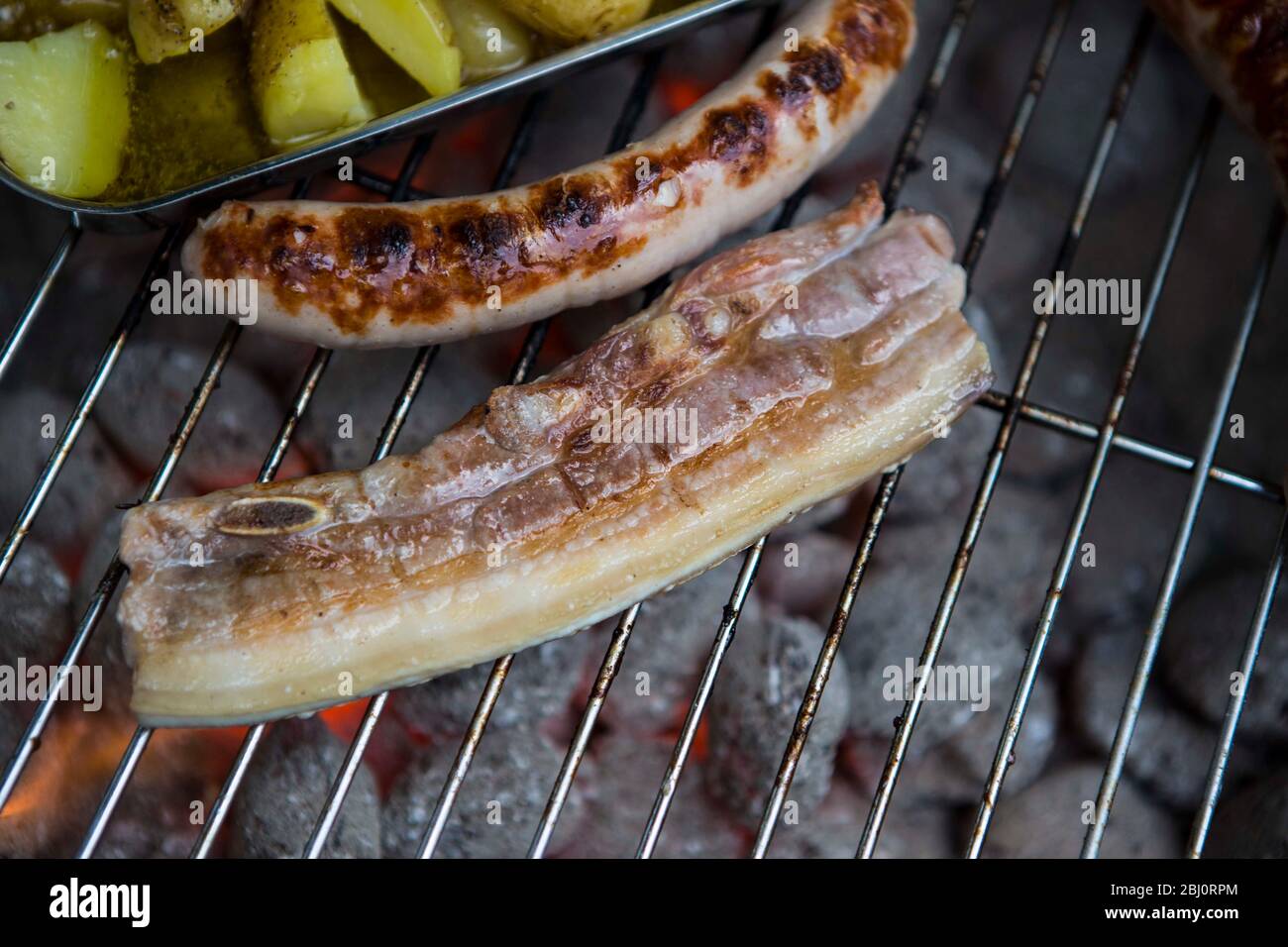 Bauchspeck vom Schwein, Bratwurst und Kartoffeln in Olivenöl mit mediterranen Kräutern auf einem Holzkohlegrill. Bauchspeck und Würstchen zählen immer Stock Photo