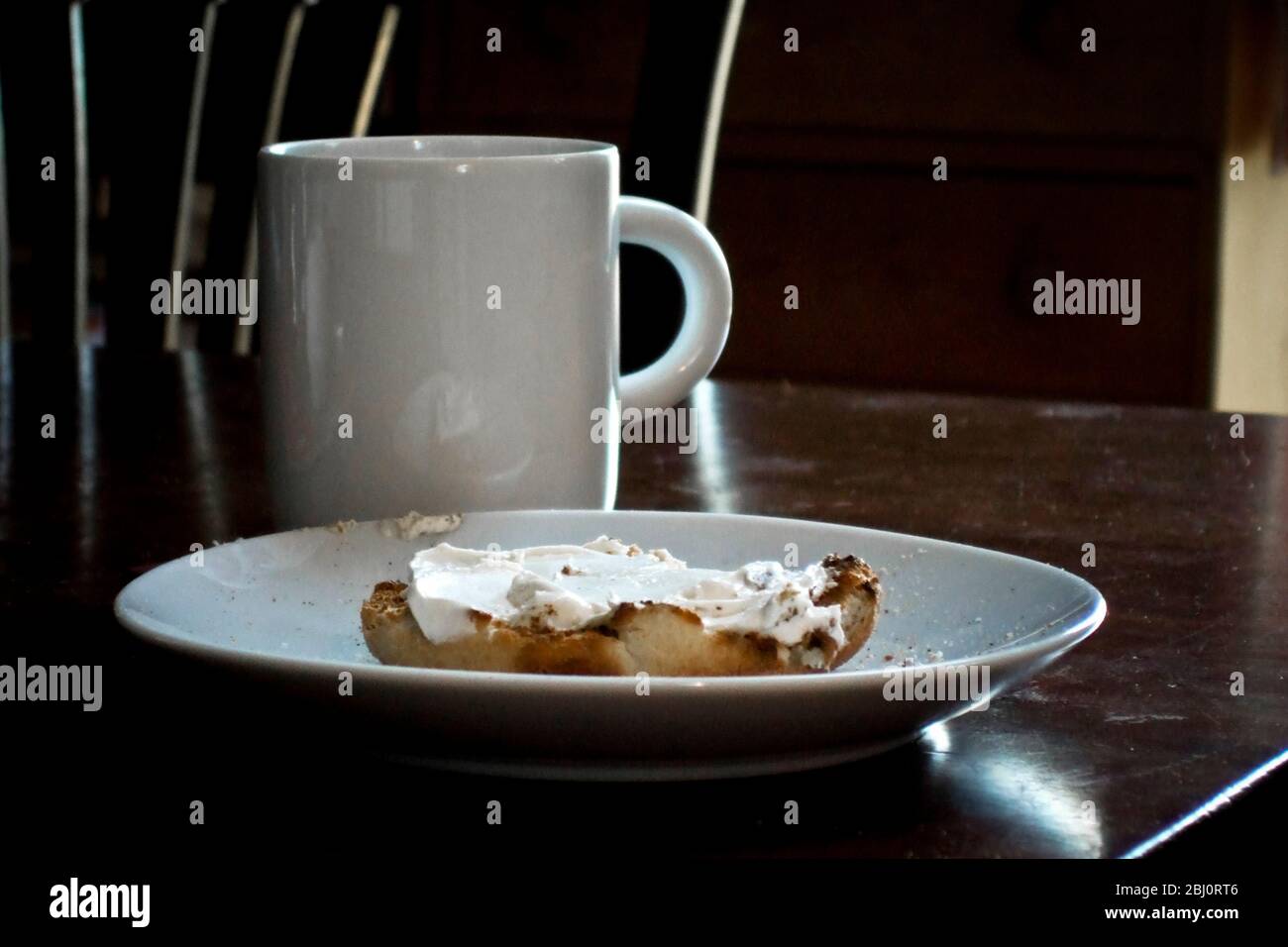 Toasted English muffin with cream cheese and bite out, with mug of coffee on polished table - Stock Photo