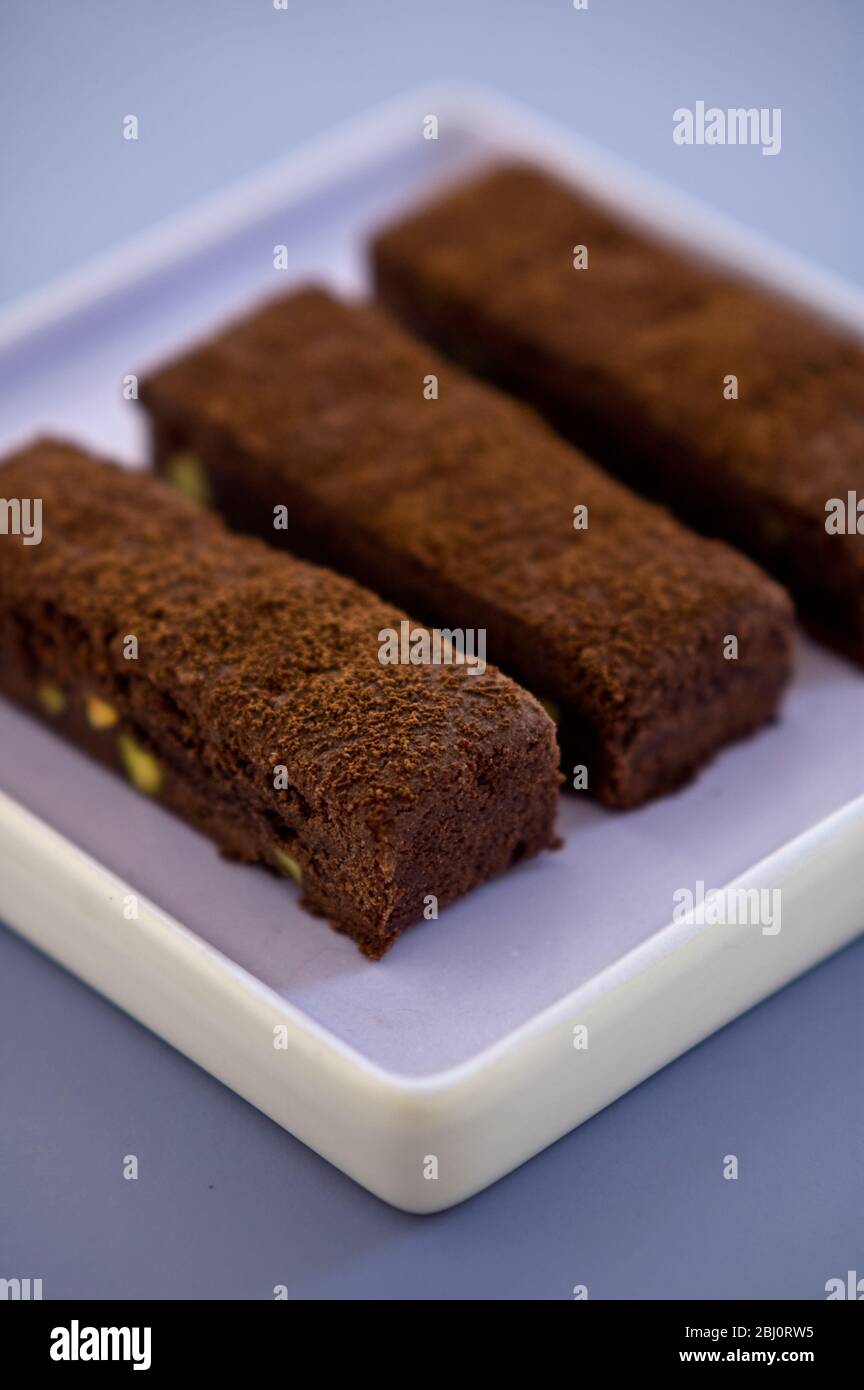 Three chocolate tray bake slices dusted with cocoa powder on square mauve plate - Stock Photo