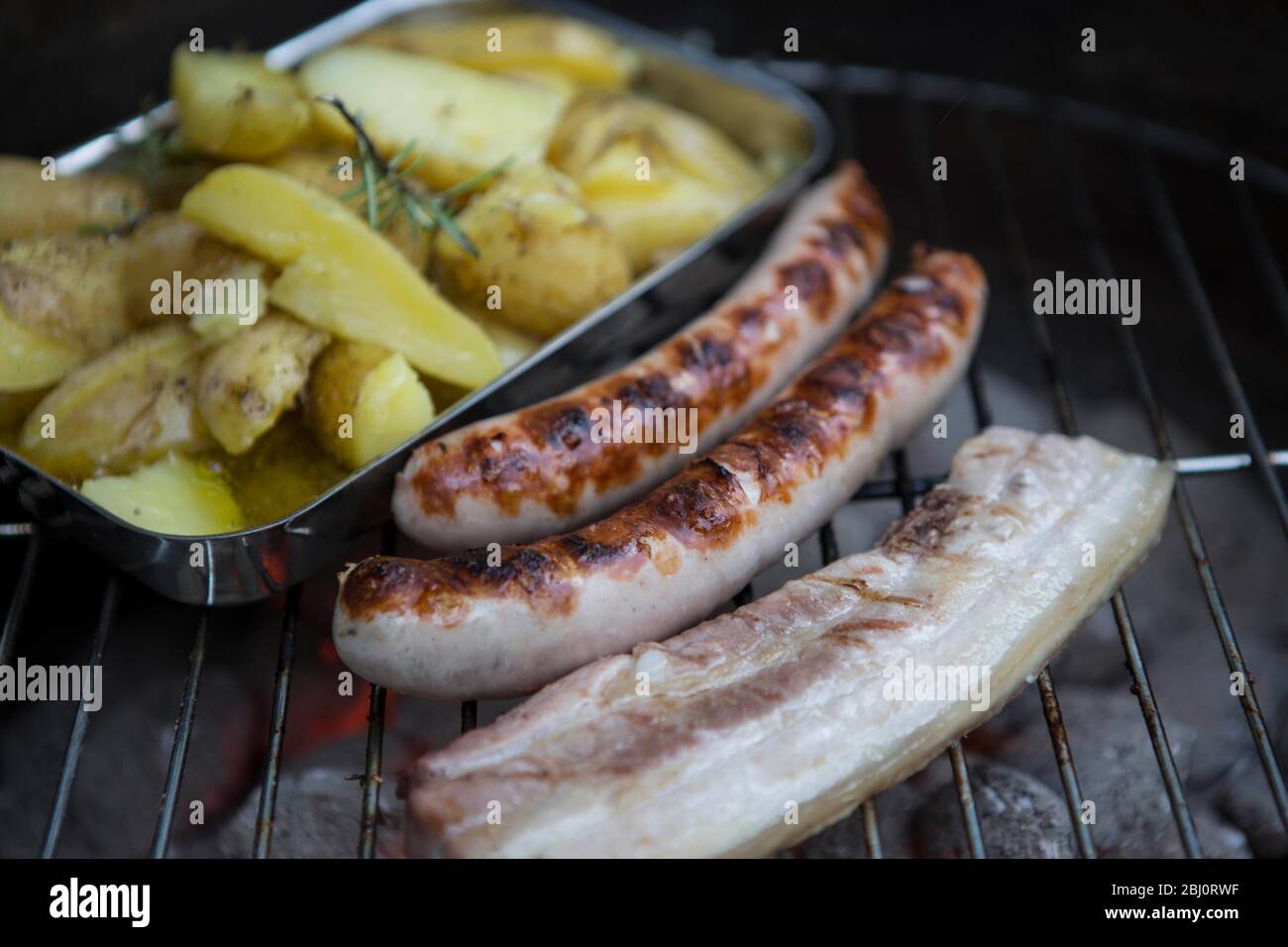 Bauchspeck vom Schwein, Bratwurst und Kartoffeln in Olivenöl mit mediterranen Kräutern auf einem Holzkohlegrill. Bauchspeck und Würstchen zählen immer Stock Photo