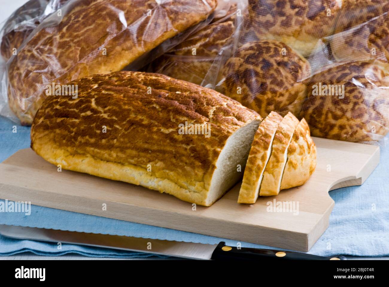Tiger' bread being sliced on wooden board with more packaged bread behind - Stock Photo