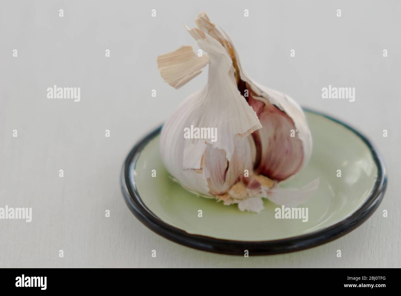 Opened head of garlic on small green enamel saucer against white background - Stock Photo