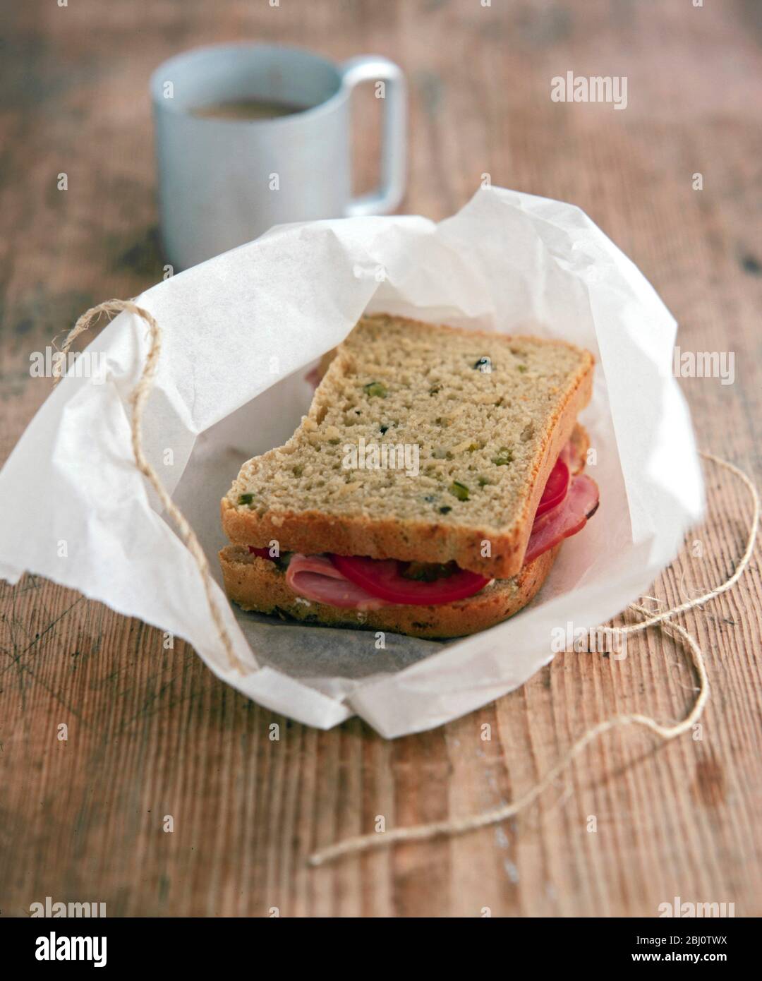 Brown bread sandwich with tomato and ham unwrapped from parcel of greaseproof paper tied with string - Stock Photo