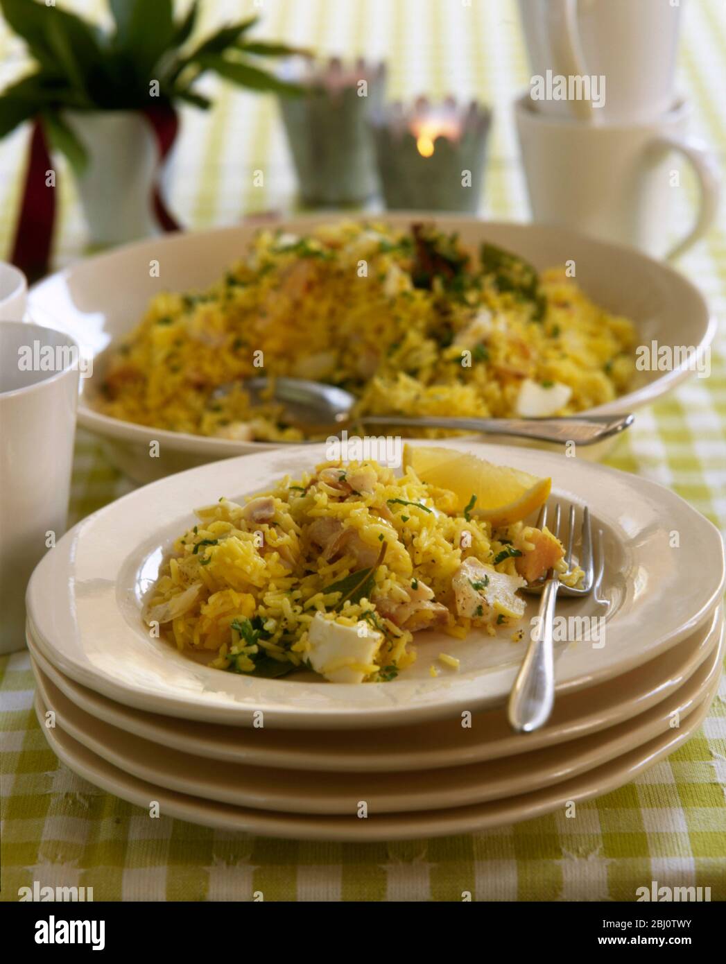 Brunch party dish of kedgeree served in dish on stack of plates with mugs and candles - Stock Photo