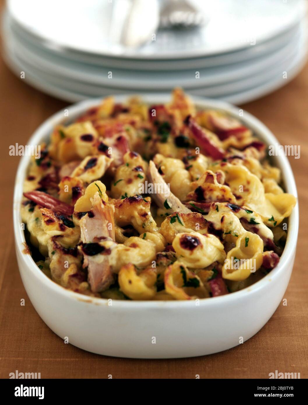 Pasta and turkey bake to use up Christmas leftovers in oven dish with pile of white plates - Stock Photo