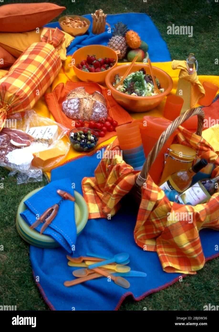 Brightly coloured picnic accessories arranged on grass, with basket of wine bottles, cheeses and salads - Stock Photo
