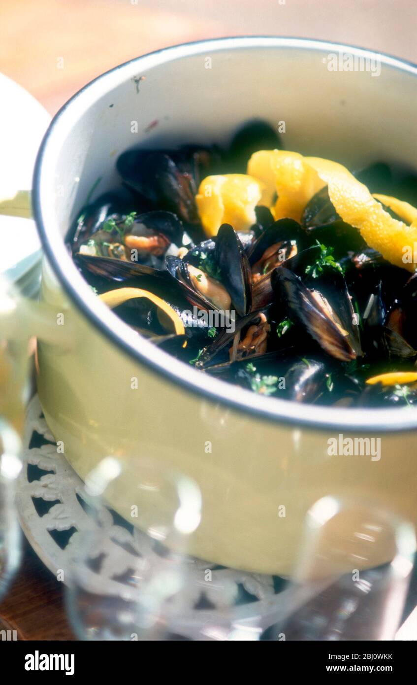 Moules mariniere served in shells with lemon rind, outdoors in cream enamel cooking pot, with wine glasses around - Stock Photo