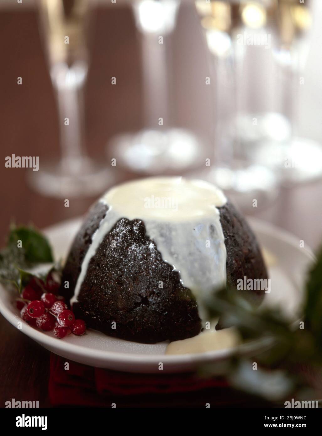 Traditional British Christmas pudding with thin cream or custard poured over and with champagne glasses in background - Stock Photo