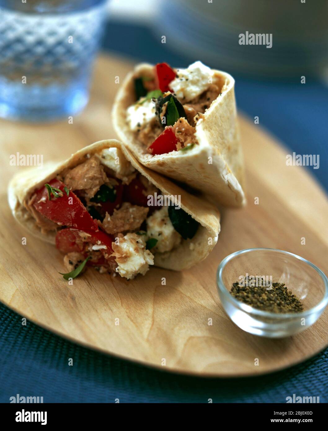 Pitta bread cut into pockets stuffed with salad of tomatoes, tuna and feta on wooden board with small glass bowl of crushed black pepper - Stock Photo