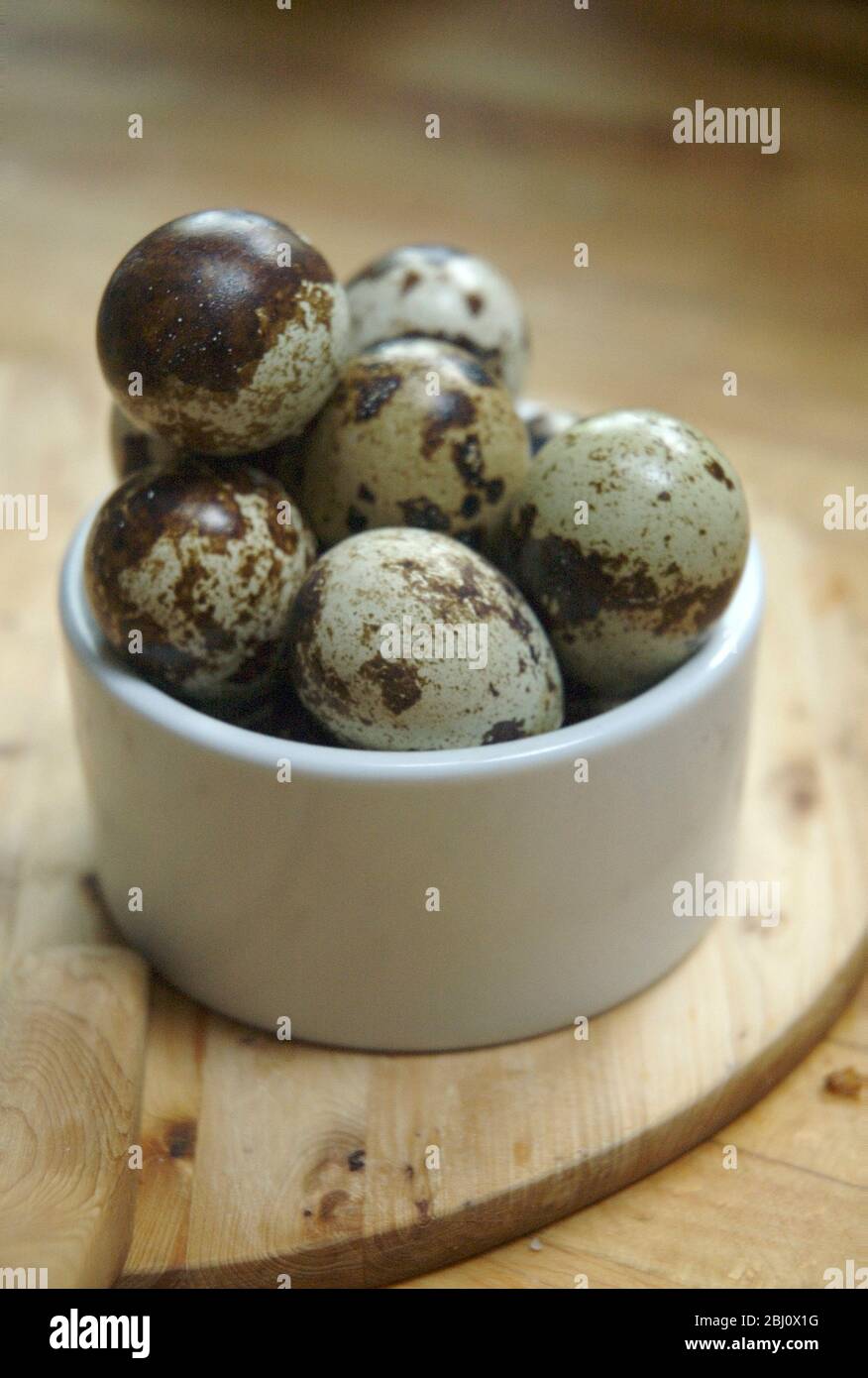 A dozen quails' eggs piled high in small ceramic white pot on wooden boards - Stock Photo