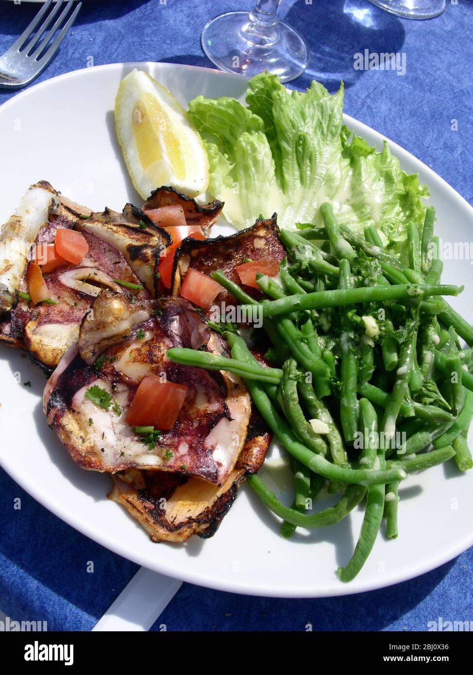Salad of grilled squid with diced tomato, green beans and lettuce with lemon wedgeon white oplate at outdoor restaurant by he sea in Menton, south of Stock Photo
