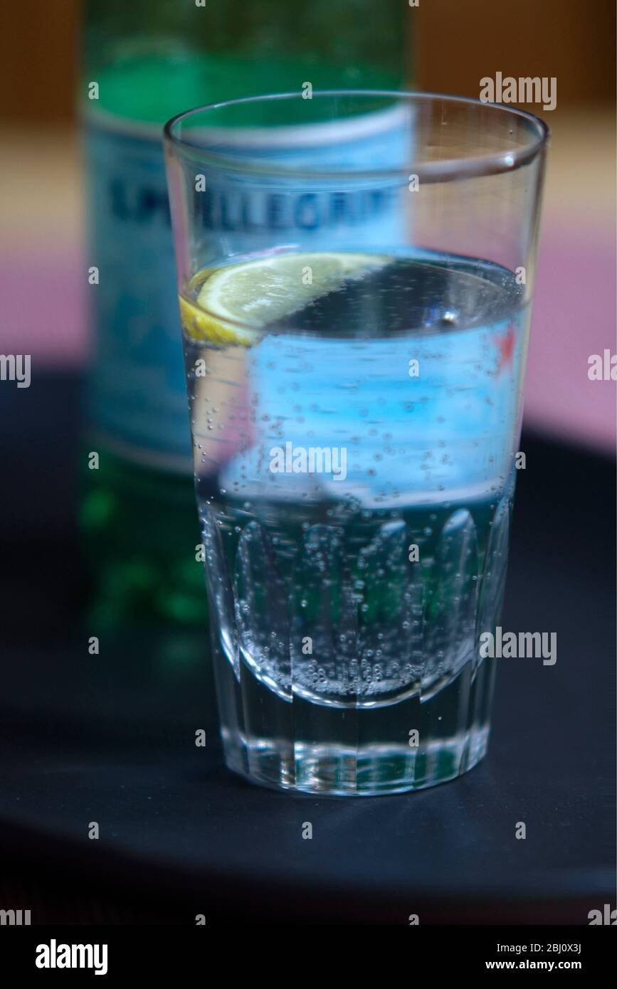 Glass of lightly sparkling Pellegrino, Italian mineral water, wirh small wedge of lemon on dark tray - Stock Photo