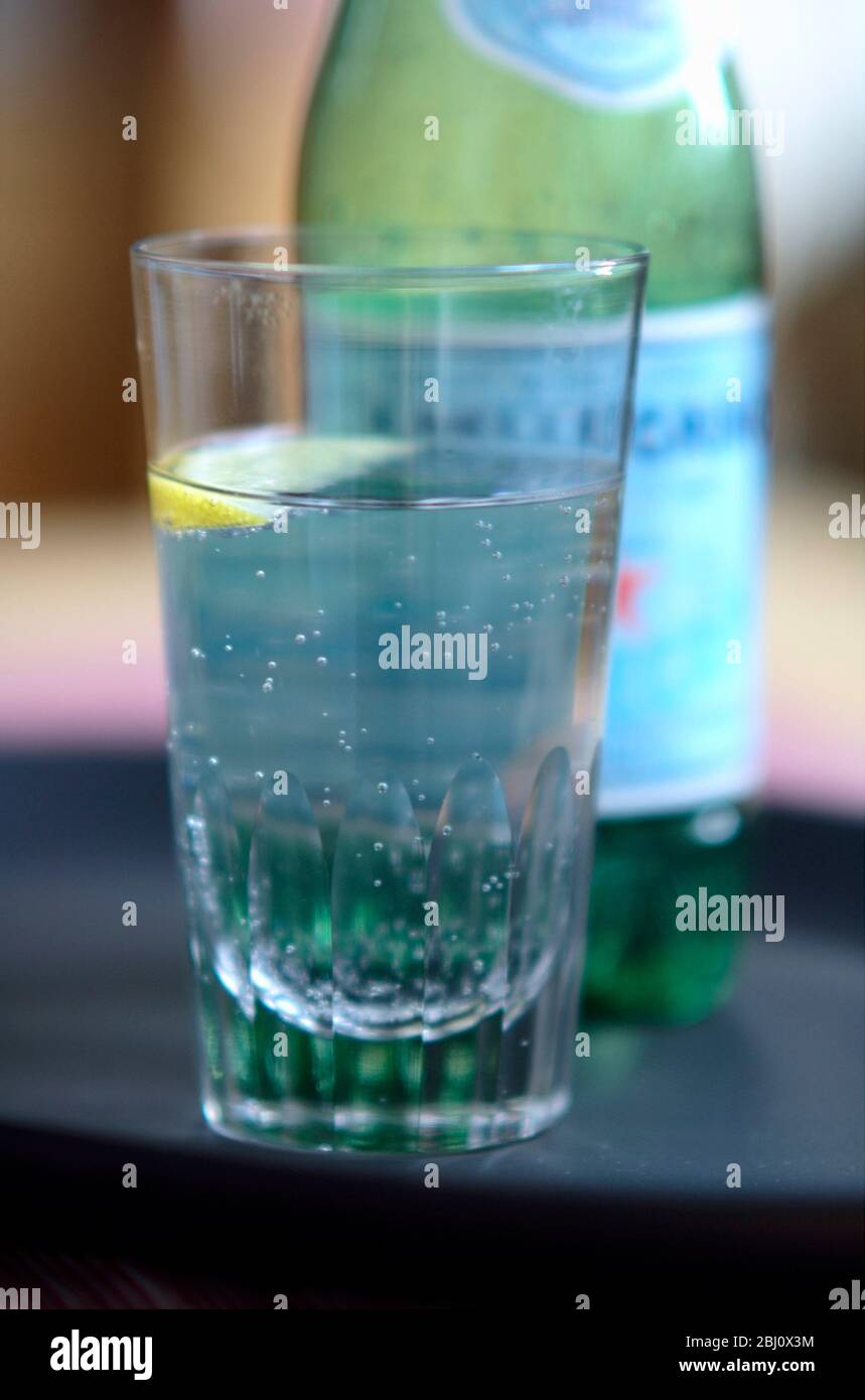 Glass of lightly sparkling Pellegrino, Italian mineral water, wirh small wedge of lemon on dark tray - Stock Photo