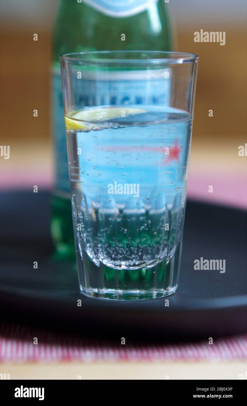 Glass of lightly sparkling Pellegrino, Italian mineral water, wirh small wedge of lemon on dark tray - Stock Photo