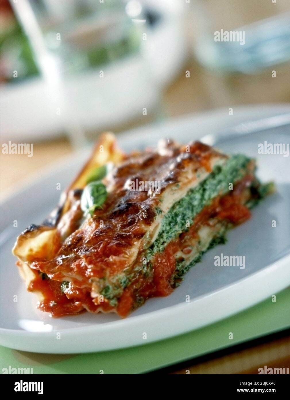 Vegetarian lasagne with spinach and tomato filling layers on white plate on green base with glasses and salad in bckground - Stock Photo