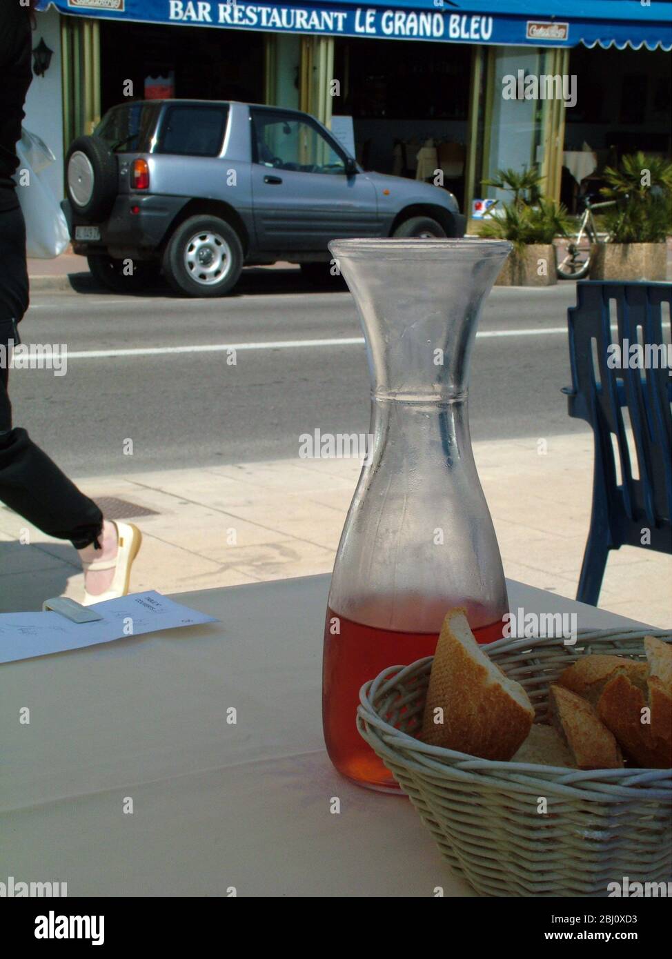 Carafe of rose wine and basket of bread on shaded table of pavement restaurant in Menton, south of France, with passer by. - Stock Photo