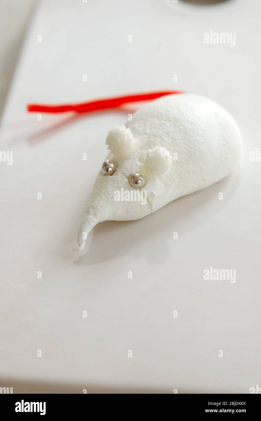 Little mouse or shrew made of meringue with silver balls for eyes and red licorice for a tail on white porcelain board - Stock Photo
