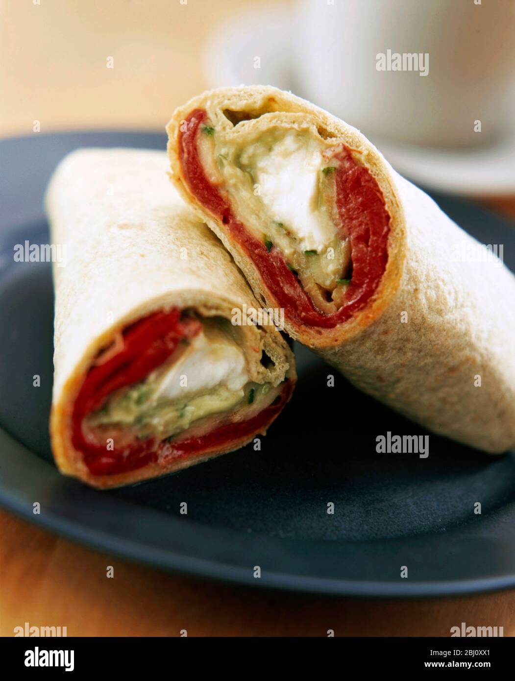 Wrap sandwich cut in two arranged on black plate with cup and saucer behind - Stock Photo