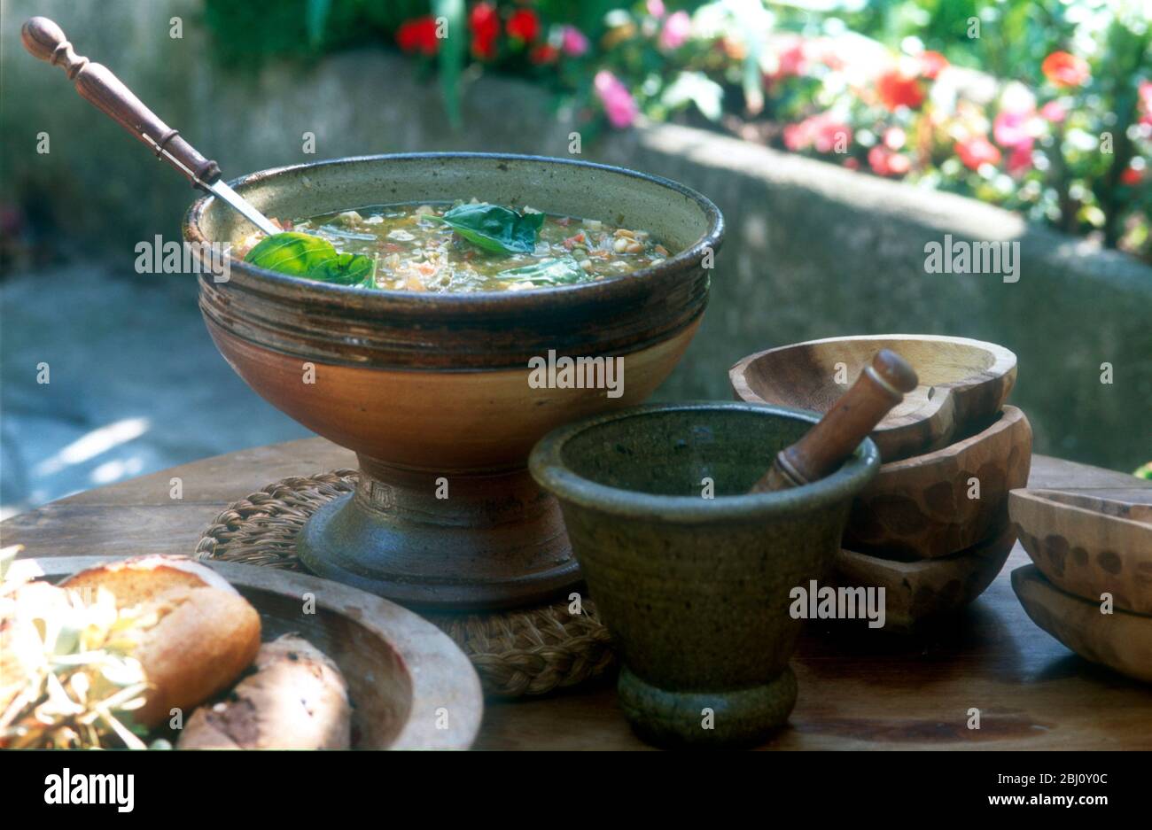 Light vegetable soup with pesto in soup tureen with bowls, bread and pesto in pestle with mortar - Stock Photo