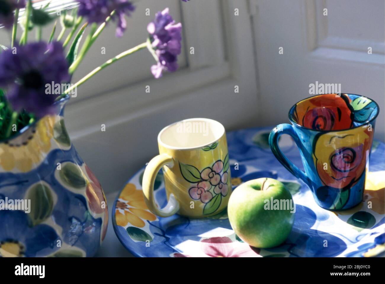 Collection of brightly painted ceramics, mugs, jug and plate, with green appole and scabious flowers in sunny window corner - Stock Photo