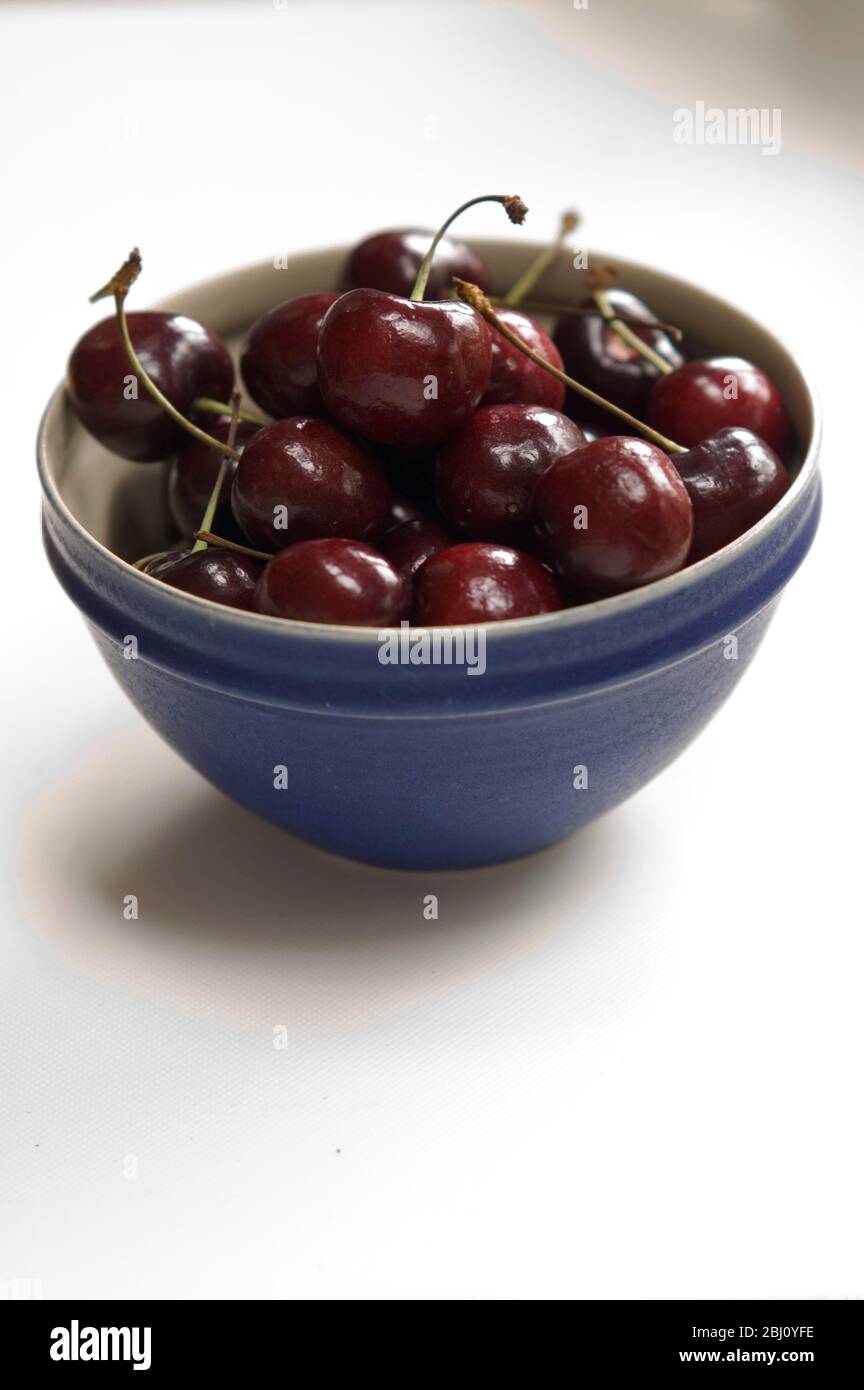 A small blue ceramic bowl of sweet red cherries on white surface - Stock Photo