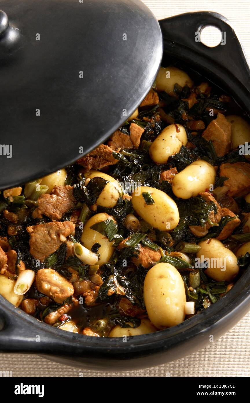 Black earthenware casserole with dish of pork spinach and potatoes with soy sauce - Stock Photo