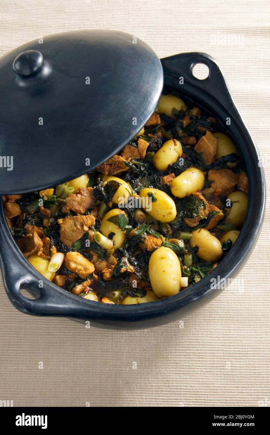 Black earthenware casserole with dish of pork spinach and potatoes with soy sauce - Stock Photo
